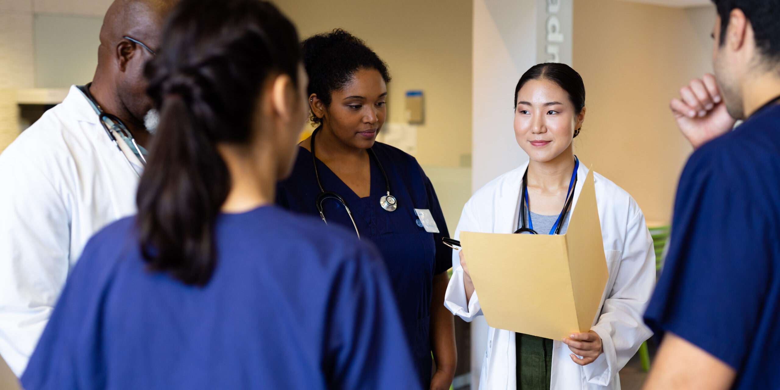 Smiling asian female doctor with folder talking with diverse colleagues in hospital corridor. Hospital, medical and healthcare services.