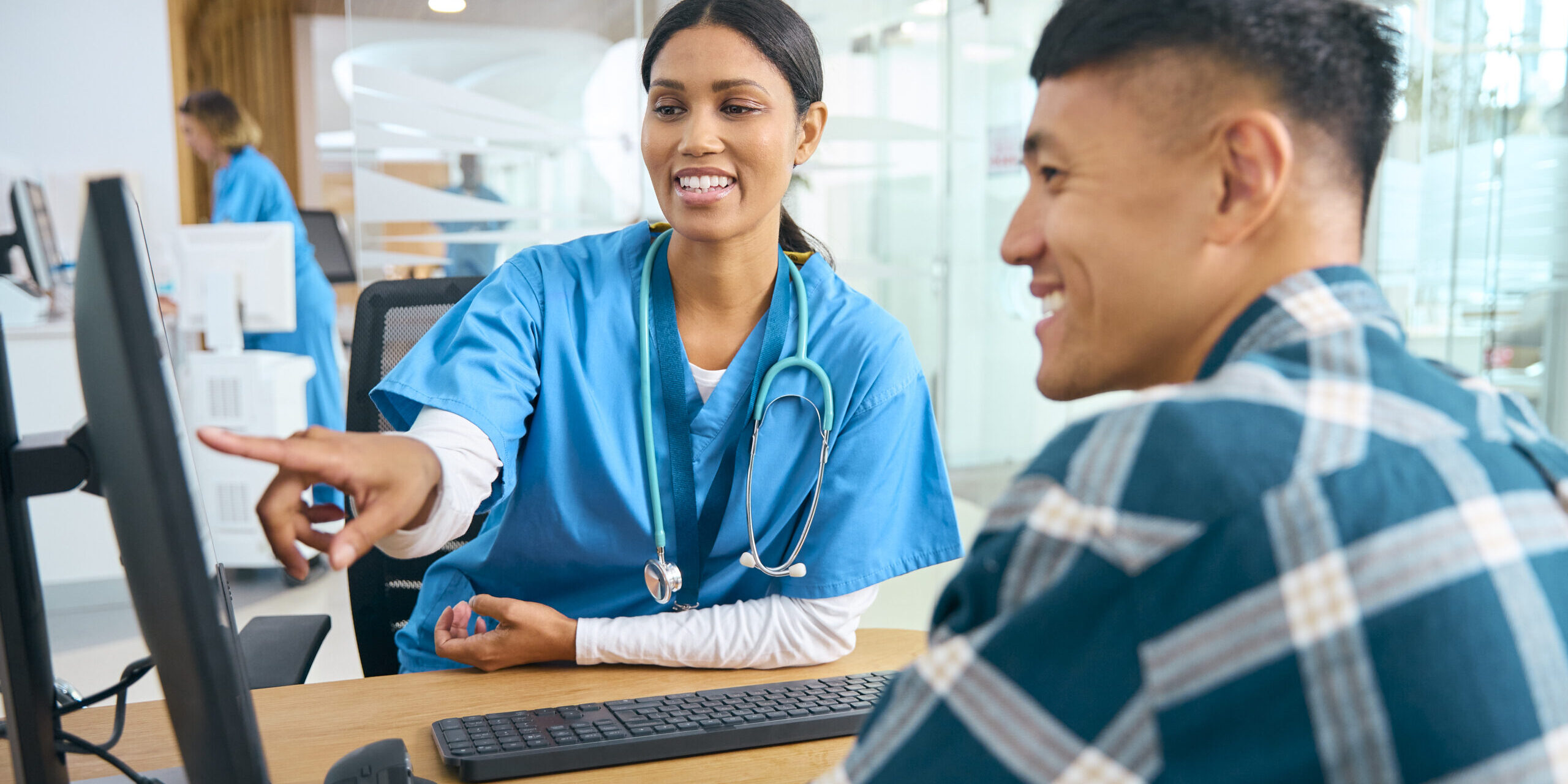 Female Nurse Or Doctor Wearing Scrubs At Hospital Appointment With Male Patient Looking At Computer